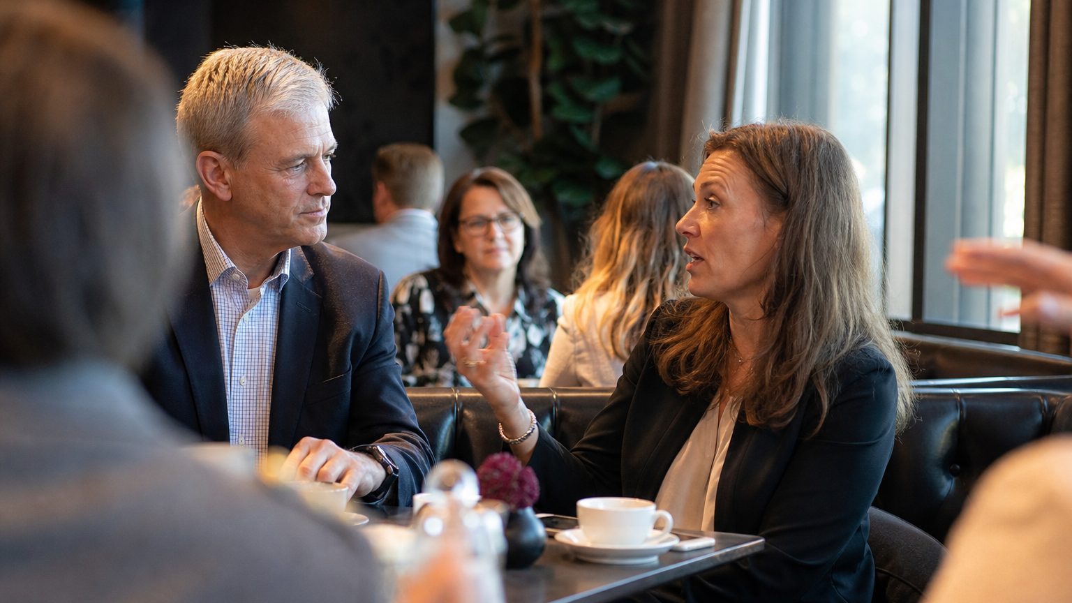 A CFO and event director sit across a restaurant table, one gesturing toward the other mid-sentence, coffee cups between them, afternoon light through a window behind them.
