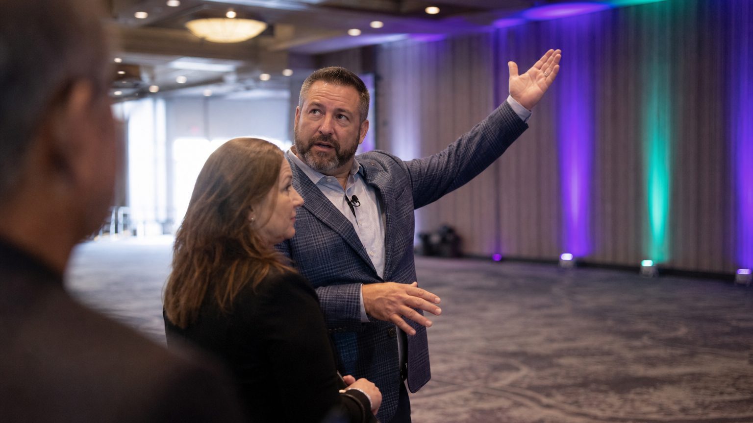A venue coordinator walks an event planner through a large conference breakout room, one arm gesturing toward the ceiling rigging while the other points toward a far wall, both mid-stride on the bare carpeted floor before setup begins.