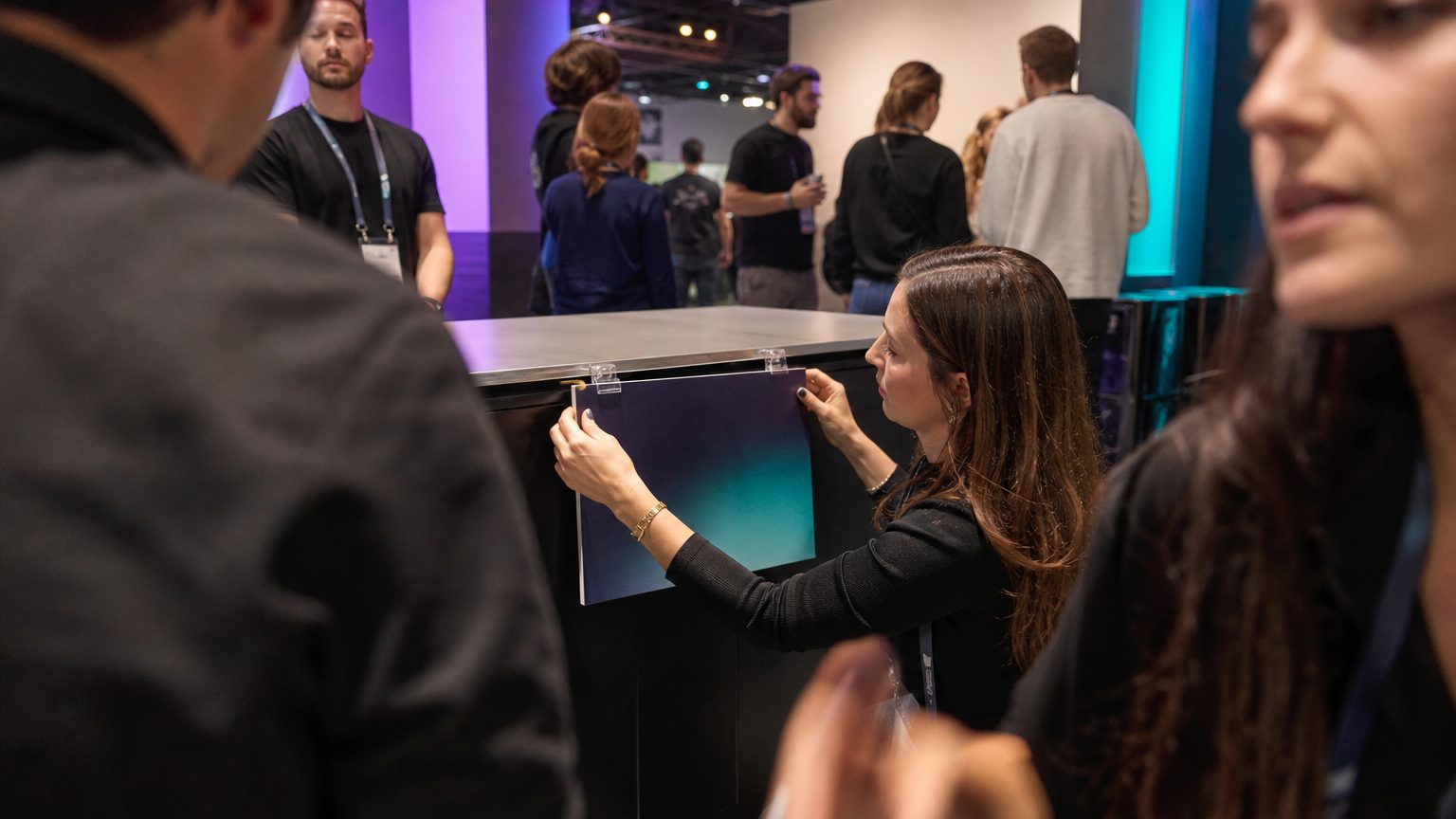 A sponsor representative clips a branded sign to the front edge of a booth counter on a busy show floor, crouching slightly to align it straight, exhibitors and attendees moving through the background behind them.