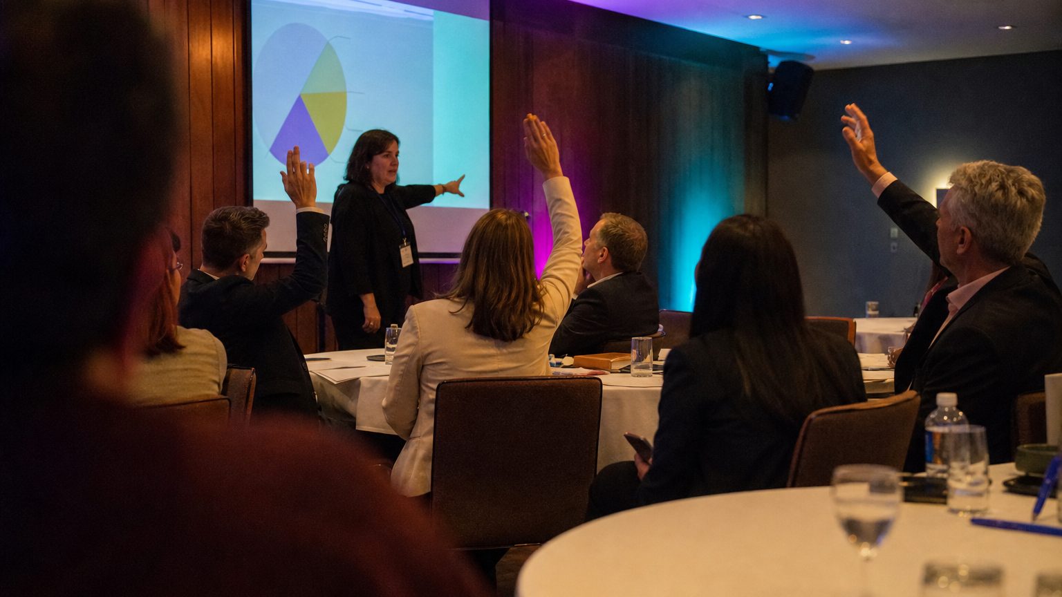 A facilitator stands at the front of a breakout session room pointing to a projected chart on a screen, while six attendees seated at round tables raise hands or lean forward to respond.