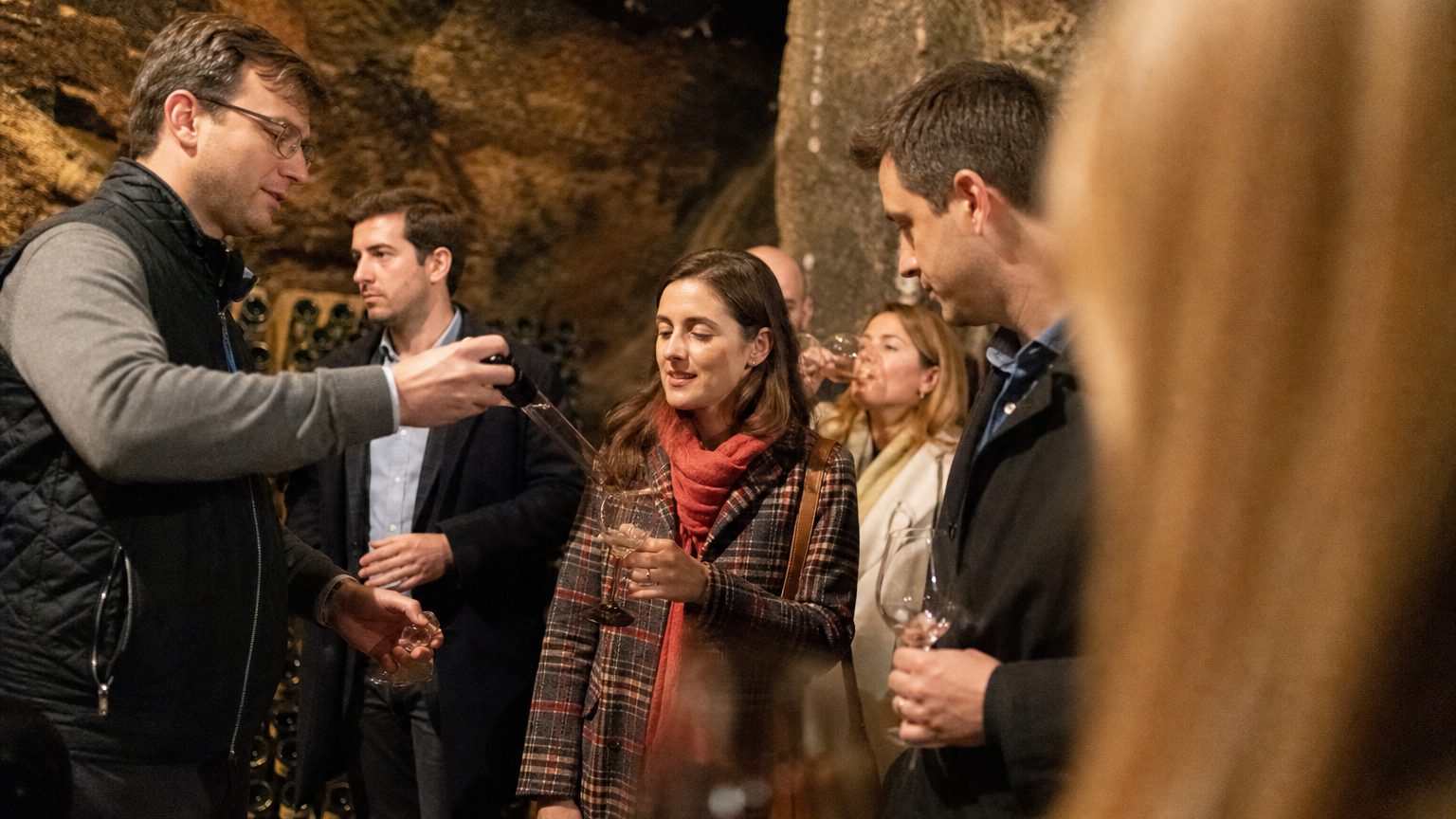 A sommelier pours a barrel sample directly into a guest's glass inside a dimly lit stone cave, the rough-hewn walls and wooden racks of aging bottles visible behind them.