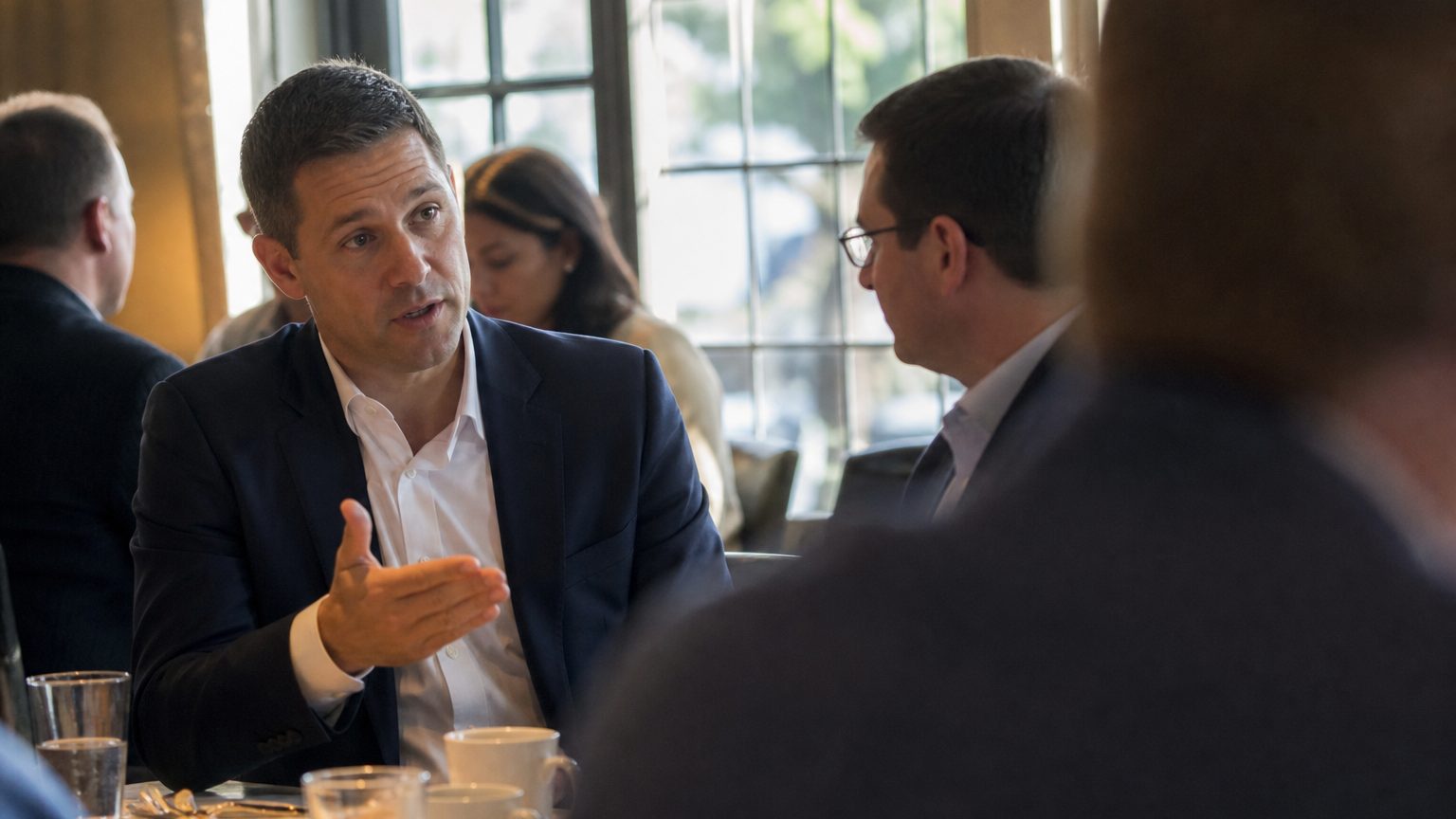 A CFO and a finance director sit across from each other at a restaurant table, one gesturing with an open hand toward the other mid-conversation, coffee cups between them, afternoon light through a window behind them.