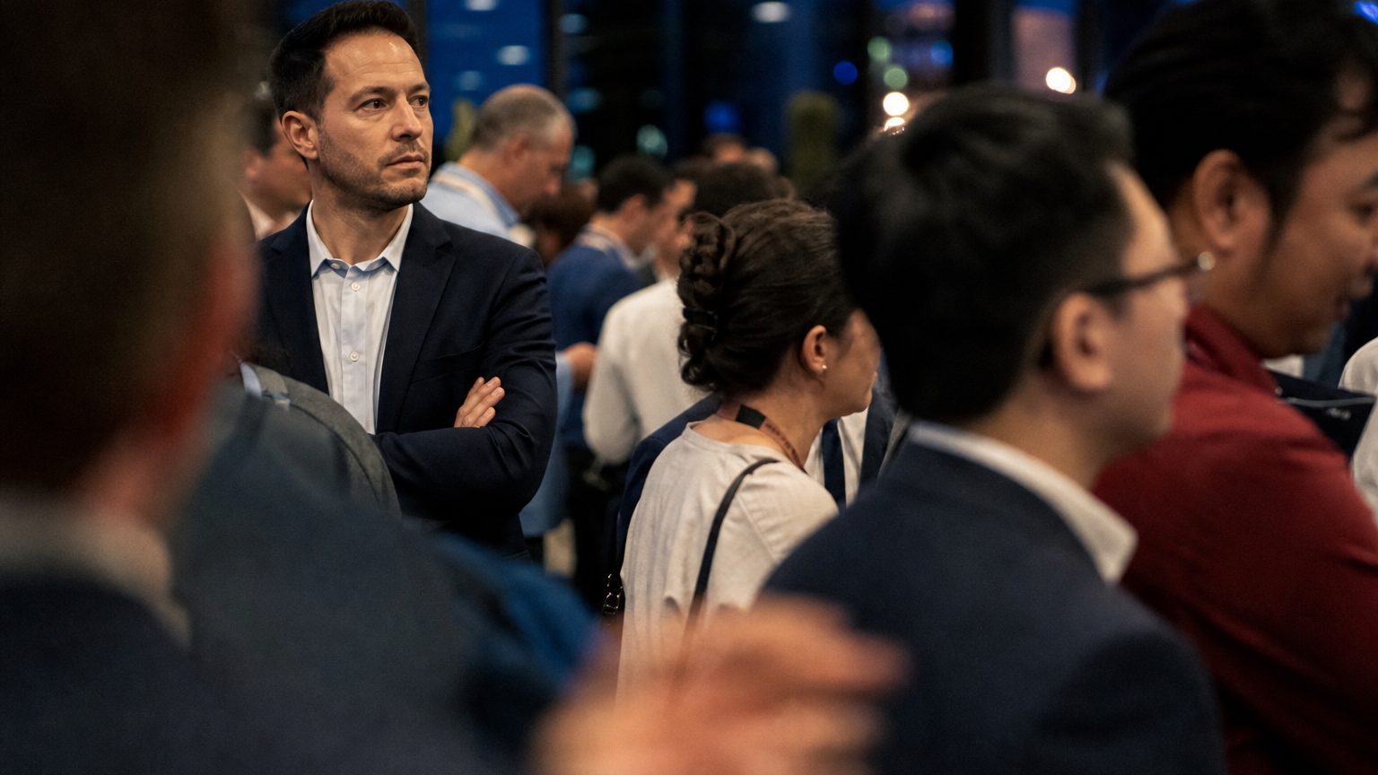 A sales rep at a trade show booth stands with arms crossed, eyes drifting past a cluster of low-value leads queuing at the counter, attention fixed on a rival booth across the aisle where a larger commission opportunity waits.