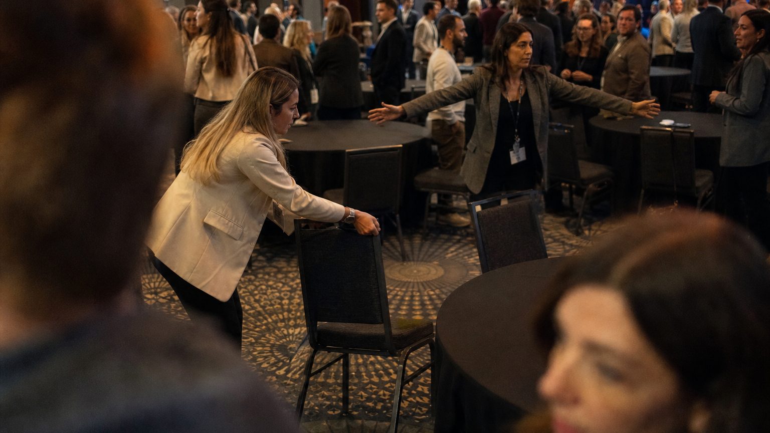 A facilitator rearranges round tables and chairs across a carpeted conference floor while a colleague measures the gap between them with outstretched arms.