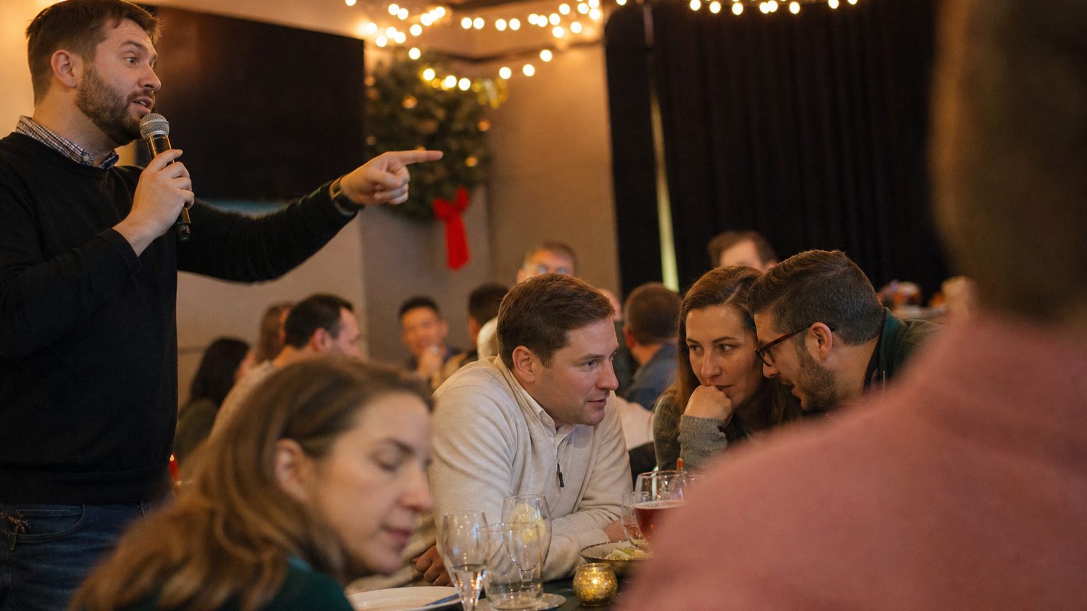 A game show host with a microphone points toward a table of colleagues leaning in to whisper an answer at a holiday team event