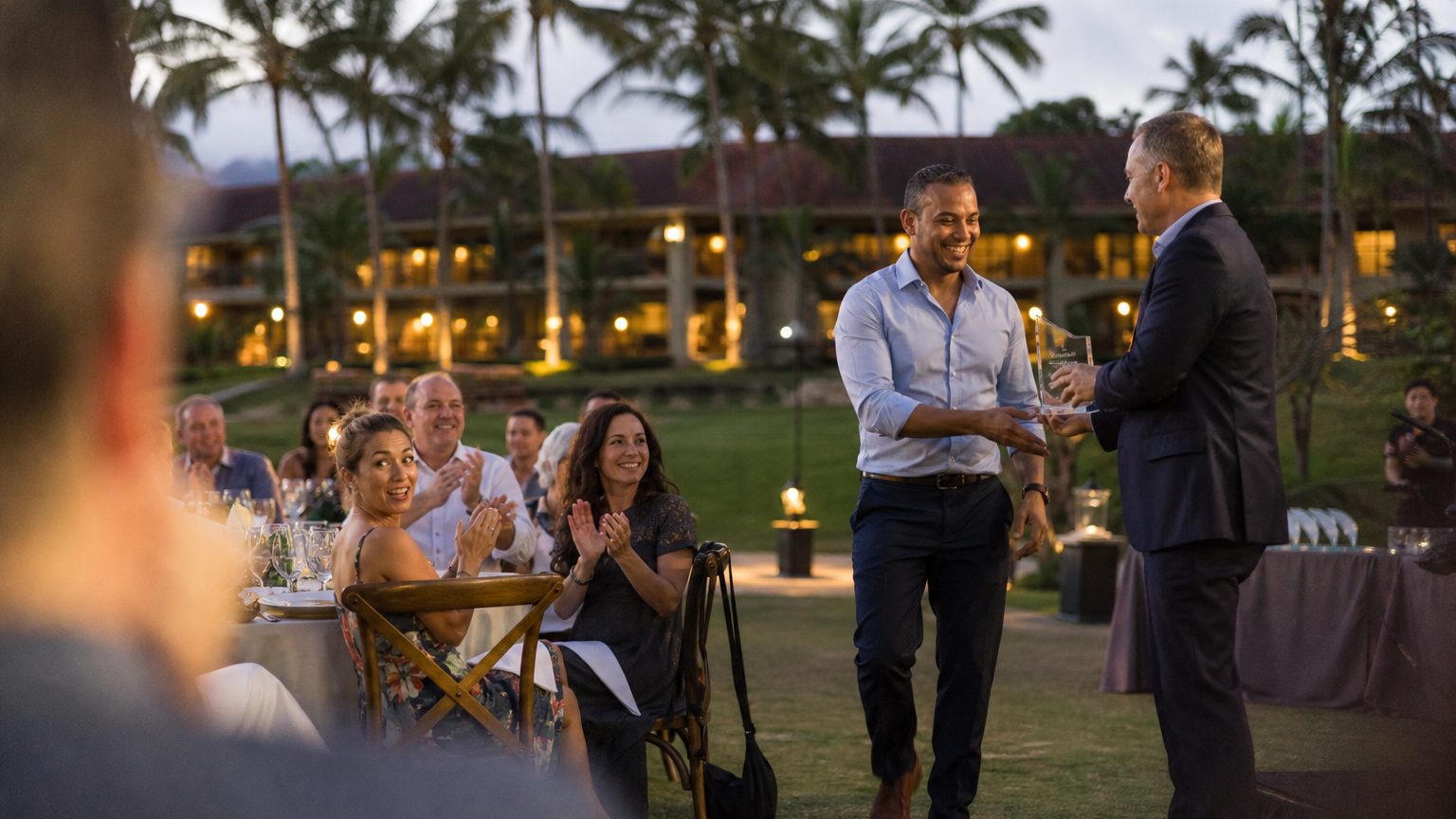 A sales rep walks up to accept a glass award from an executive at an outdoor awards dinner as colleagues applaud