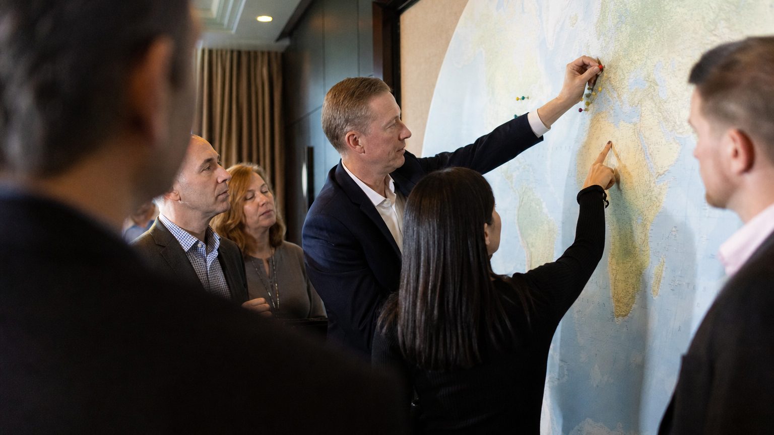 A travel manager stands at a large wall-mounted world map in a hotel conference room, pressing a colour-coded pin into a coastal region while a colleague points to a different cluster of pins already marking three other shortlisted locations.