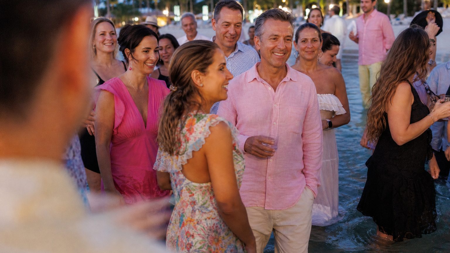 A group of attendees in resort wear wade into shallow turquoise water at the edge of a white-sand Caribbean beach, late-afternoon sun casting long shadows behind them.
