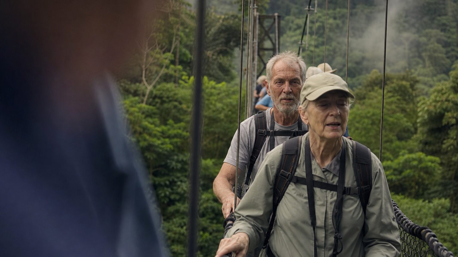 Two attendees in hiking gear cross a hanging suspension bridge above a dense Costa Rican rainforest canopy, late morning light filtering through mist rising from the valley below.