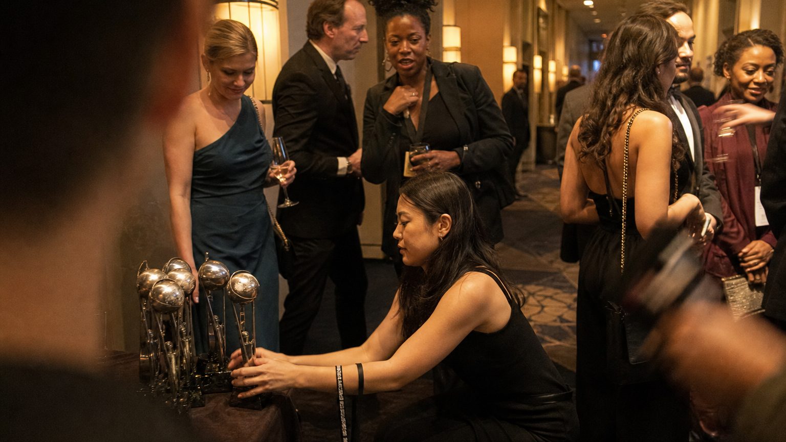 A prop stylist crouches beside a cluster of award trophies on a draped table in a hotel corridor, arranging them by height while a lanyard bearing the event name hangs from her wrist.